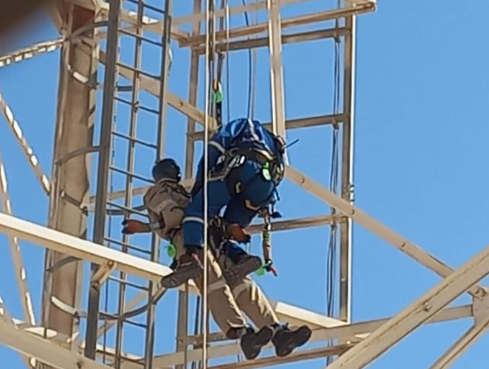 A rescue worker in a blue jumpsuit and safety harness assists an incapacitated person in tan overalls suspended by ropes on a white metal lattice tower under a clear blue sky.